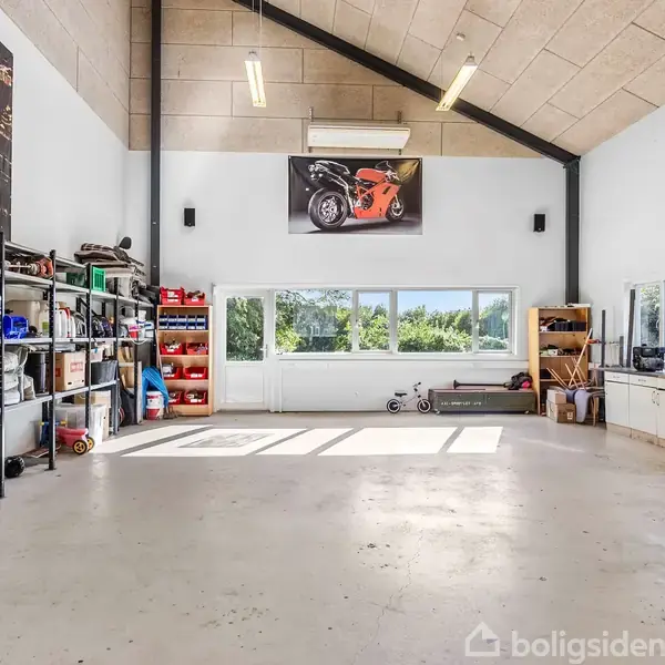 Et stort værksted med højt loft, indeholder reoler med opbevaringskasser til venstre, arbejdsborde til højre og vinduer mod haven. Plakat af en motorcykel hænger på væggen.