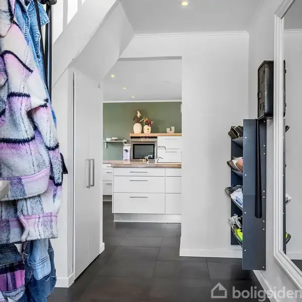 En entre med hvide vægge og sorte gulvfliser. Til venstre hænger farverige jakker. Modellen fortsætter ind mod et køkken med hvide skabe og en grøn væg i baggrunden.