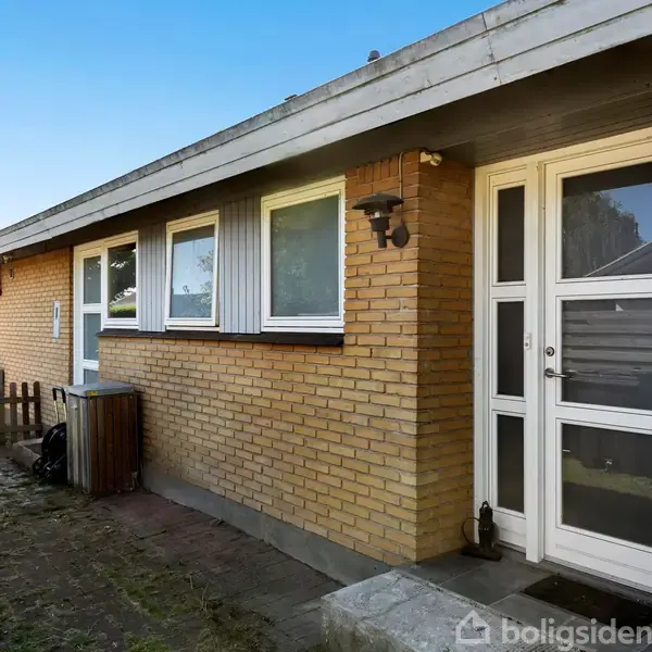 Rødt murstenshus med hvid hoveddør og tre vinduer, beliggende i en have med græsplæne og et lille hegn. Lampen ved døren tilføjer lys til indgangen.