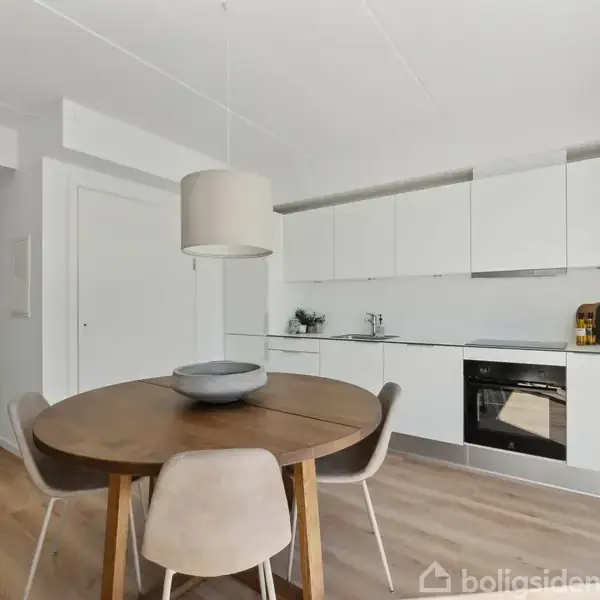 Et moderne køkken med hvide skabe og indbygget ovn. Rundt spisebord med grå stole står i forgrunden, mens en stor lampe hænger over bordet.