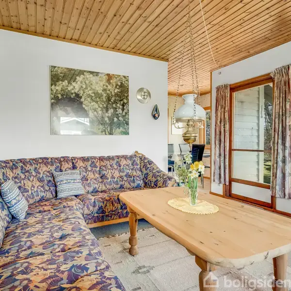 En stor blomstret sofa står i en stue med træloft, ved siden af et træbord med en vase med blomster. Ved siden af er der en stor glasdør med udsigt til haven.