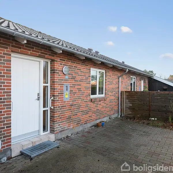 Rødt murstenshus med hvid dør står i solskin. Nummer 1A er ved hoveddøren. Foran er et flisebelagt indgangsparti, omkranset af en lav træhegn.