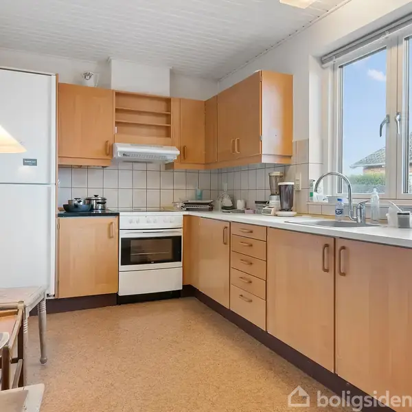 Et køkken med træskabe, hvidt køleskab og komfur. Det står i et lyst rum med fliser over køkkenbordet og vindue, hvorigennem man kan se et tag og himmel.