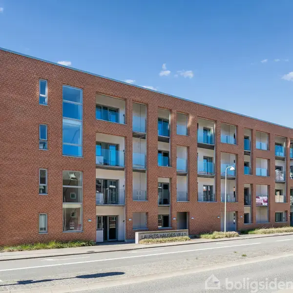 En murstensbygning med store vinduer og altaner står langs en vej med fortov. Skiltet foran bygningen lyder "Laurits Walthersvej 29". Solskin og blå himmel i baggrunden.