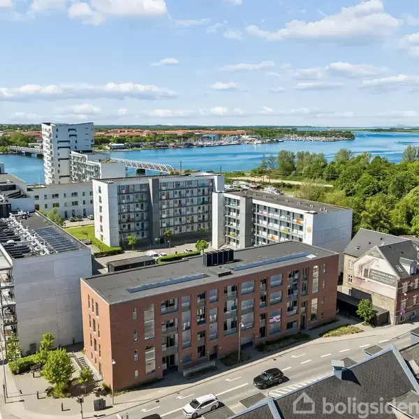 Flere moderne lejlighedsbygninger står langs en byvej, mens en stor sø med grønne områder strækker sig i baggrunden under en blå, skyet himmel.