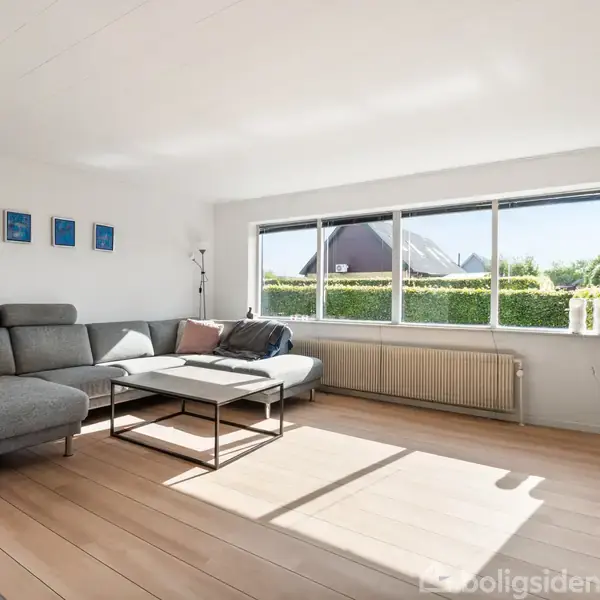 En grå hjørnesofa med puder står i en lys stue. Foran sofaen er der et sofabord. Store vinduer giver udsigt til en grøn hæk og en naboens tag.
