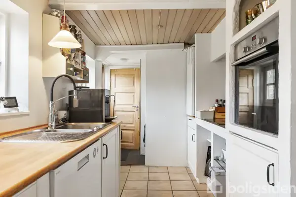 Et køkken viser en vask med en kaffemaskine ved siden af. Der er en ovn indbygget i væggen, og konteksten er en lys, funktionel indretning med træloft og flisegulv.