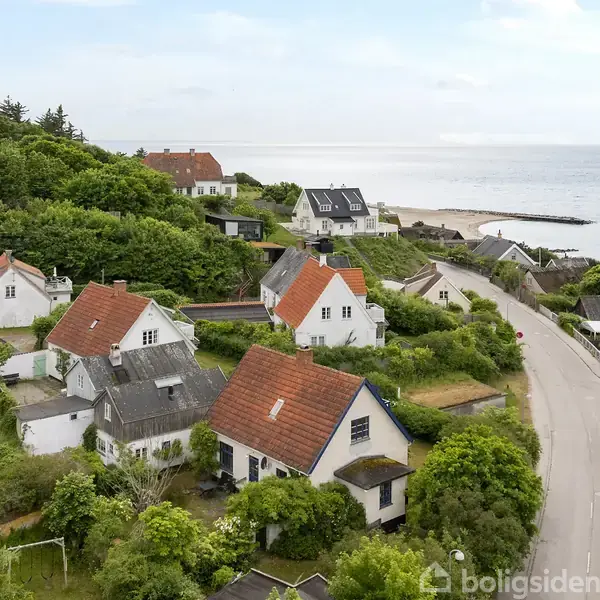 Huse med røde tage står stille i et frodigt, kuperet landskab ved siden af en snoet vej, tæt på en kystlinje og havudsigt, omkranset af grønne træer og buske.