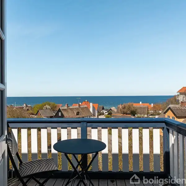 Balkon med sort cafébord og stol står i forgrunden. Bagved ses tage fra huse og et blåt hav med klar himmel.