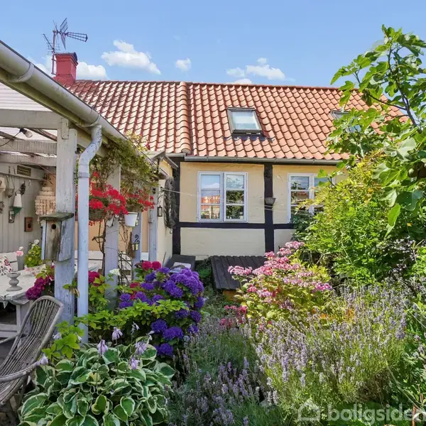 Et bindingsværkshus står i en farverig have med blomster, mens en overdækket terrasse til venstre har træmøbler og hængende planter. Det er solrigt og fredfyldt.