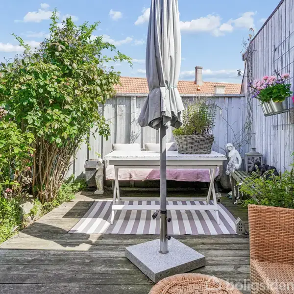 Terrasse med parasol i midten, omgivet af træmøbler og grønne planter. Et trægulv med tæppe strækker sig foran en hvidmalet trævæg med blomster i potter.