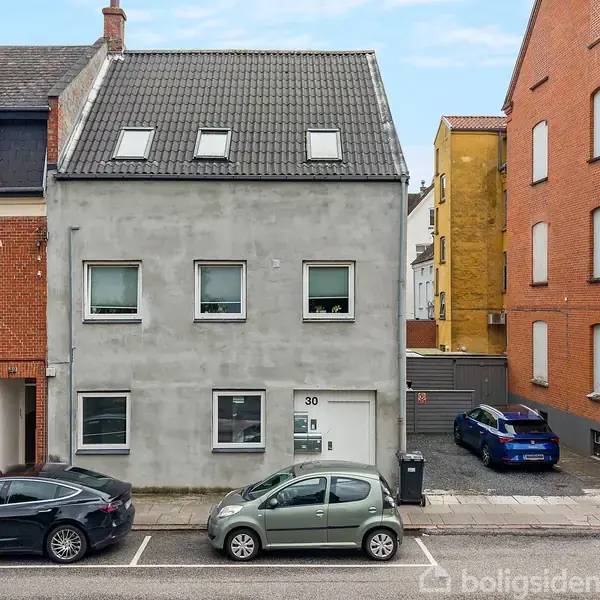 Gråt, toetagers hus med skråt tag og to ovenlysvinduer, nummer 30 på facaden. Omgivet af røde murstenshuse. To parkerede biler foran på gaden.