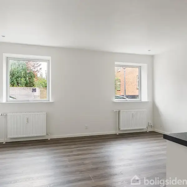 Stue med grå trægulv, tom for møbler, har to store vinduer. Radiatorer placeret under hvert vindue. Lyse vægge giver et rummeligt indtryk. Urban udsigt udenfor.