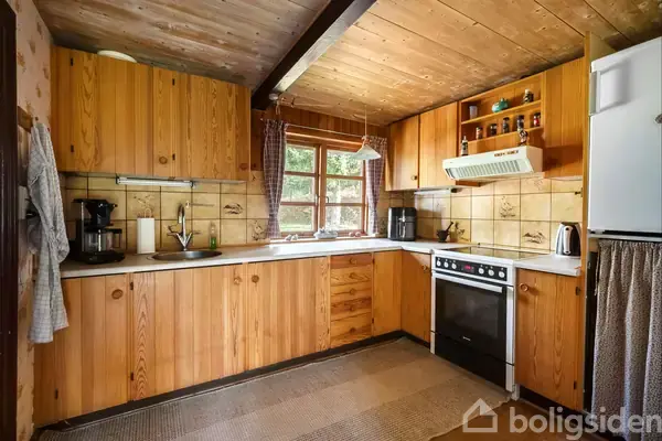 Et køkken med træskabe og en ovn. En kaffemaskine og andre køkkenredskaber står på bordpladen med flisebelagt væg. Et vindue giver udsigt til grønt udenfor.