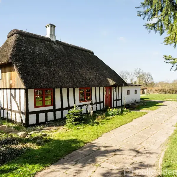 Stråtækt bindingsværkshus står stille i solbelyst have med græsplæne og træer. Flisebelagt sti fører til indgang. Rødmalede vinduer er synlige på husets facade.