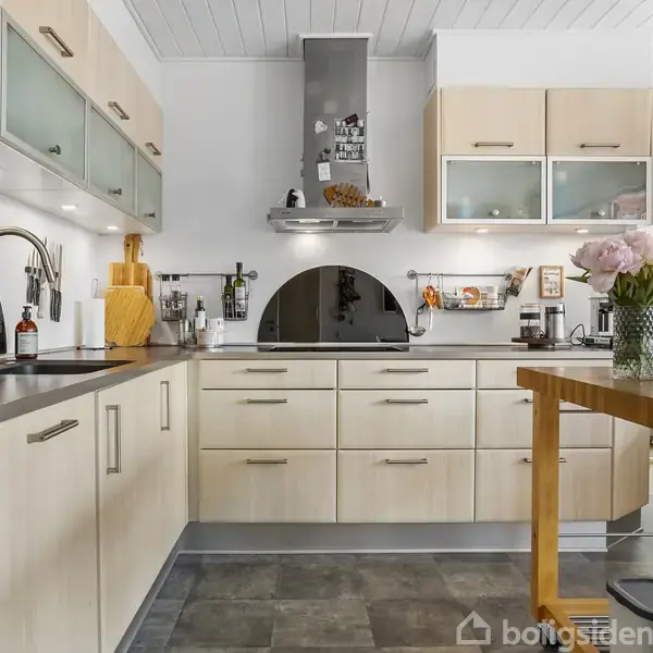 Et moderne køkken med lyse træskabe og en mørk bordplade. Kogeplade under emhætte, blomster i vase på et træbord. Klokke og lamper i baggrunden.