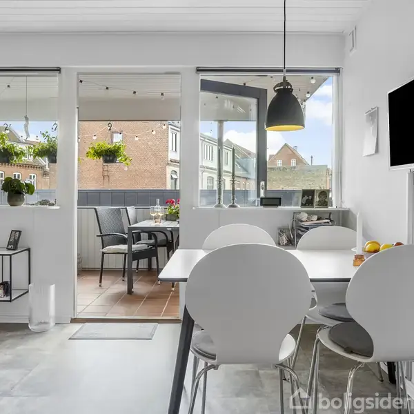 Spisebord med hvide stole står i et lyst køkken. Udenfor ses en altan med planter og en cafébordopsætning. Store vinduer lader lys ind, og der hænger en lampe over bordet.