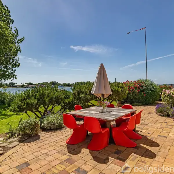 Et udendørs spisebord med røde stole står på en terrasse. Bordet har en parasol i midten. Terrassen er omgivet af grønt med udsigt over en sø og blå himmel.