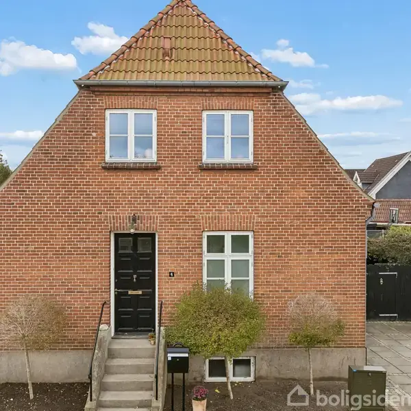 En rød murstensvilla med sort hoveddør og en trappe, flankeret af små træer, beliggende i et boligkvarter med andre huse. Huset har to vinduer på første sal.