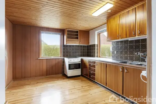 Komfur står ubrugt i køkken med træpaneler på vægge og loft. Køkkenet har hvide skabe, sort flisebagvæg og to vinduer, der giver dagslys. Gulvet er i lyst træ.