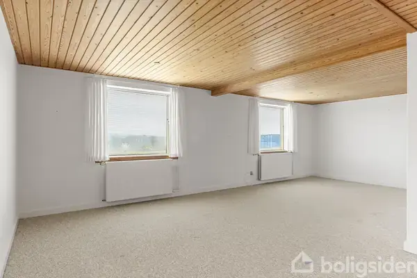 Et tomt rum med lyst gulvtæppe og træloft. To vinduer med hvide gardiner slipper lys ind. Radiatorer er placeret under hvert vindue, hvilket skaber et funktionelt boligmiljø.