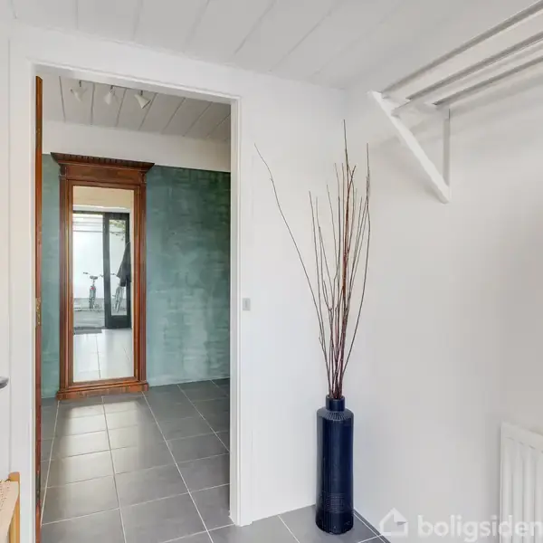 En indgang giver udsigt til en gang med spejl og grønne vægge. En høj vase med grene står til højre, og der er en knagerække over.