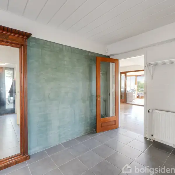Spejl med træramme står op ad en grøn væg, reflekterende lys. En åben trædør leder til et rum med trægulv, og en hylde med taske hænger over en radiator.