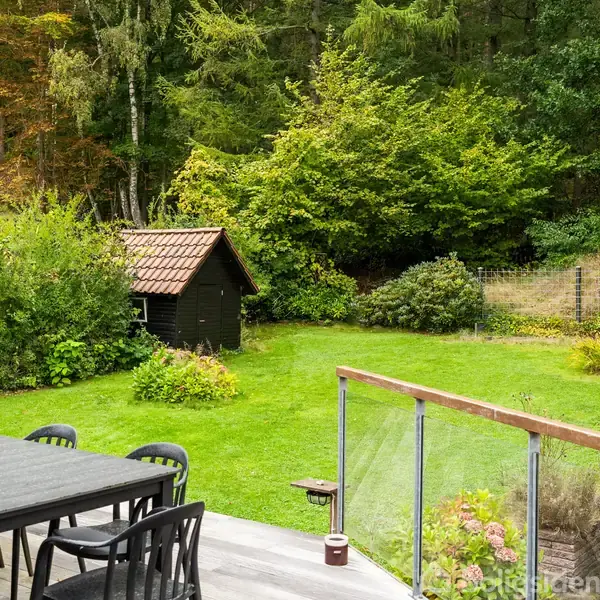 Et sort havebord med stole står på en terrasse. Terrassen har glasgelænder og åbner ud mod en grøn have med et lille skur og træer i baggrunden.