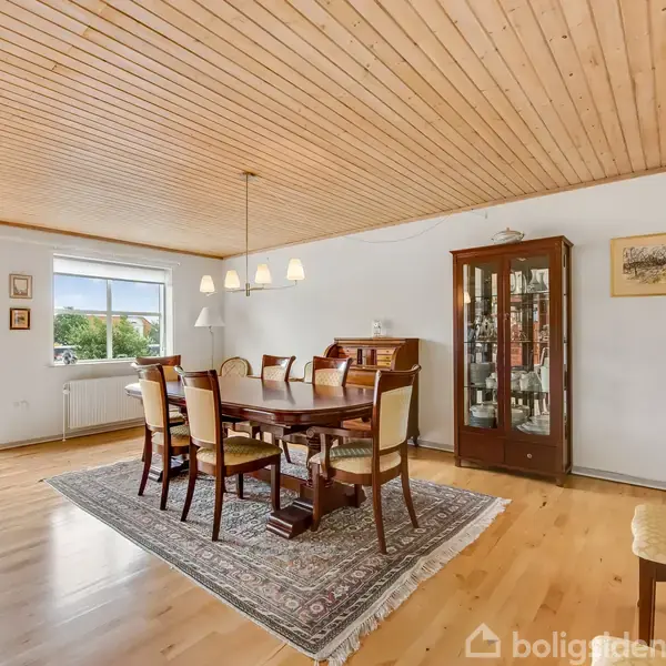 Spisebord står i midten af en spisestue, omgivet af seks stole. Et vitrineskab og en kommode står mod væggen. Store vinduer og en dør giver udsigt til en balkon.