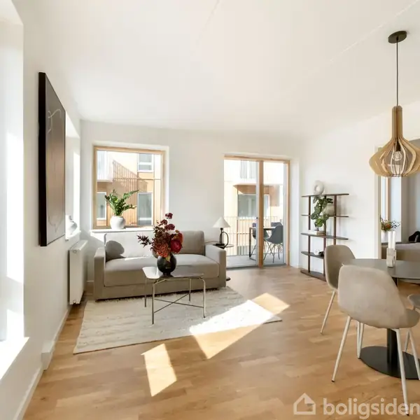 En lys stue indeholder en grå sofa med blomsterdekoration, placeret ved store vinduer. Et rundt spisebord med fire stole står under en moderne lampe. Værelse med trægulv og hvide vægge.