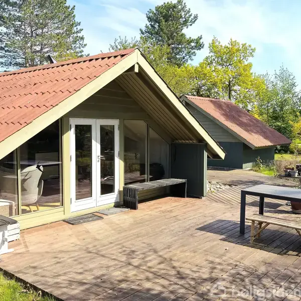 Hyggeligt sommerhus med rødt tag og grøn facade står på en træterrasse. Indendørs ses stole gennem glasdøre. En anden bygning ses i baggrunden omgivet af træer.