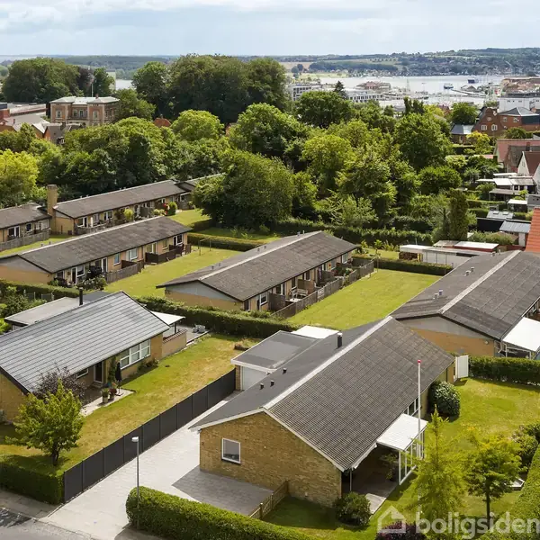 Rækkehuse med flade tage ligger i et grønt boligområde. Omkring dem er velplejede haver og buske. I baggrunden kan man se en by og en havn.