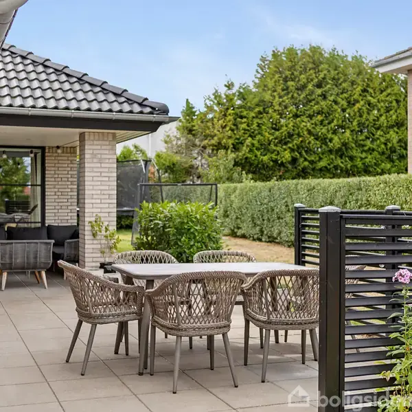 Udemøbler står på en flisebelagt terrasse ved siden af et hus med lyse mursten. Terrassen er omkranset af en sort metalafskærmning og omgivet af grønne hække og planter.