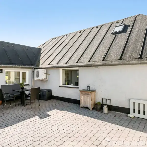 Et hvidt hus med sort tag står i sollys. På den brostensbelagte gårdsplads er der en udendørs siddegruppe. Baggrunden viser grønne marker under en klar blå himmel.
