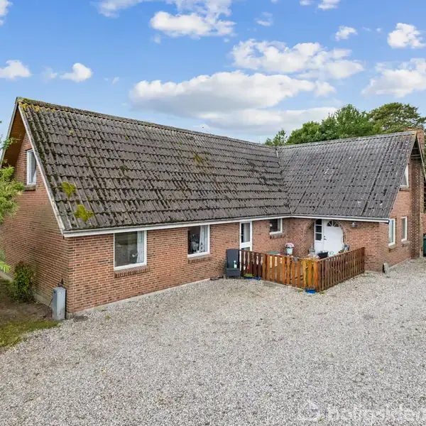 En stor brun murstensbygning med skråt tag, omgivet af en grusbelagt gårdsplads. Der er en garage til højre og træer i baggrunden under en skyfri himmel.