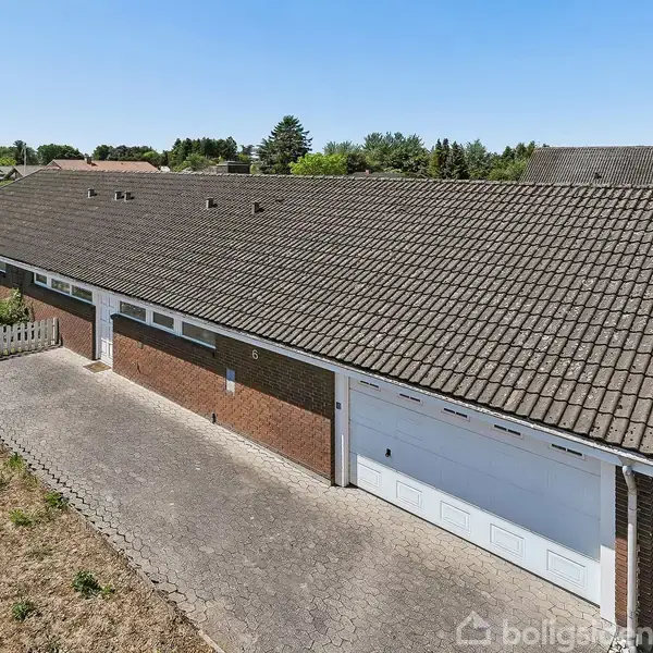 En lang, brun murstensbygning med gråt tegltag har en tilknyttet garage. Foran er der en flisebelagt indkørsel. Omgivet af græs og nogle træer i baggrunden.