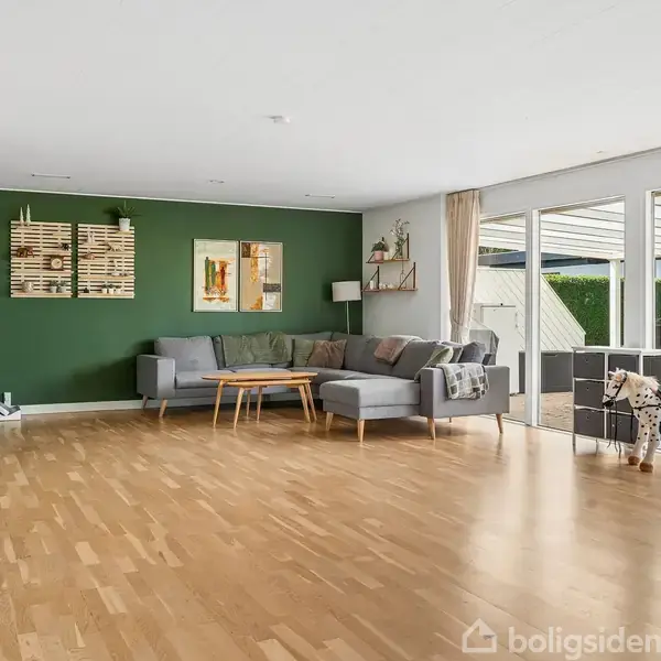 Stue med en grå hjørnesofa og sofabord mod en grøn væg, dekoreret med kunst. Store vinduer åbner ud til en terrasse med havemøbler. Trægulv og indendørs planter.