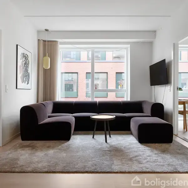 En mørk sofa står på et lyst tæppe i en stue med hvide vægge. Et lille rundt bord står foran sofaen. Store vinduer viser en moderne bygning udenfor.