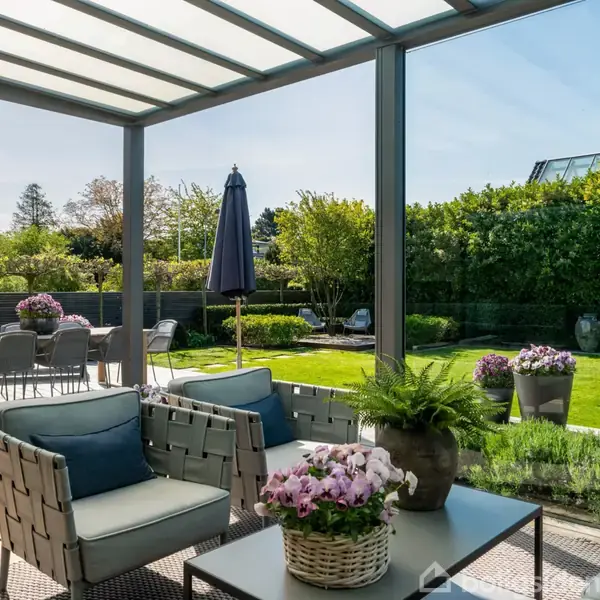 Terrassemøbler står under en overdækket pergola; i baggrunden ses en have med grøn græsplæne, blomsterkrukker og et bord med parasol. Vejret er klart, og omgivelserne er frodige.