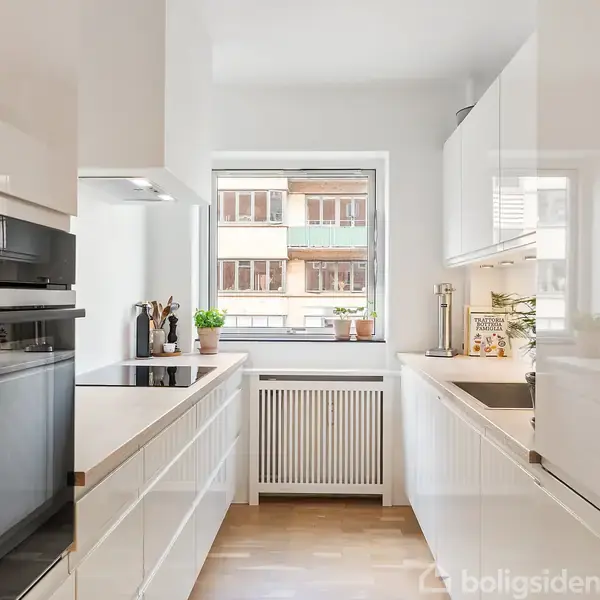 Et moderne køkken med hvide skabe og apparater. Køkkenet har en indbygget ovn, glaskeramisk kogeplade, og vindue med udsigt til en bygningsfacade. Pottedekorationer ses ved vinduet.