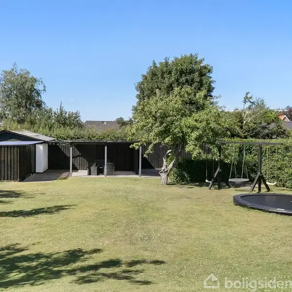 En bred græsplæne med en trampolin, omgivet af grønne buske og træer. I baggrunden ses et sort hus eller skur og en terrasse. Himlen er klar og blå.