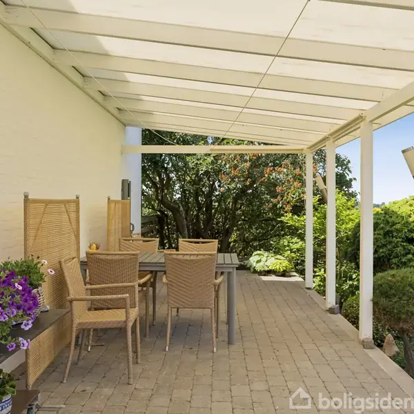 Udendørs terrasse med træbord og stole står under en hvid pergola. Der er blomster og grønne planter omkring, hvilket skaber en hyggelig atmosfære i en haveomgivelse.