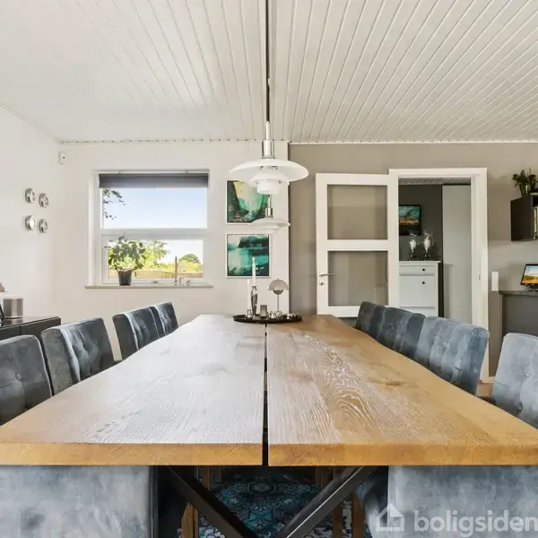 Et langt spisebord med blågrå stole står i et lyst rum. En lampe hænger over bordet, og en stor kunstmaleri dekorerer væggen. Vinduet giver udsigt til grønne områder.