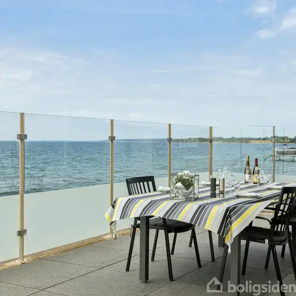 Et bord dækket med en stribet dug står på en terrasse. Der er fire sorte stole, og bordet har vinflasker og glas. Udsigten viser havet og en kystlinje.