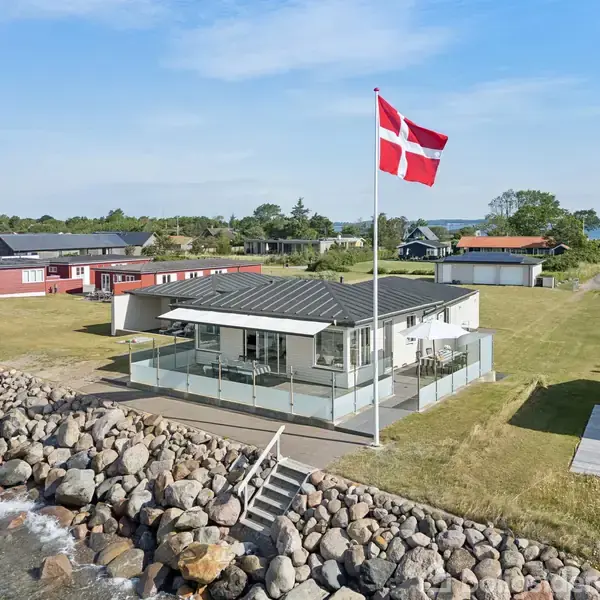 Et moderne hus med glasindhegnet terrasse står langs en kystlinje af store sten. Det danske flag vajer fra en høj flagstang i haven, omgivet af grønne græsplæner.