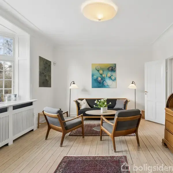 Stue med trægulv hvor to lænestole og en sofa omkranser et rundt sofabord med blomster. Maleri hænger på væggen; lys strømmer ind fra store vinduer.