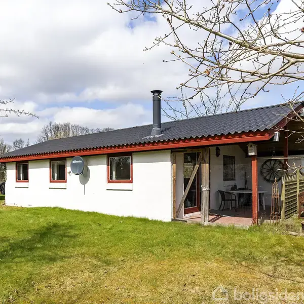 Et enkelt, hvidt hus med rødt tag og en skorsten står på en grøn græsplæne omgivet af træer. Der er terrassemøbler under en overdækket veranda. Ingen tekst bortset fra "boligsiden."