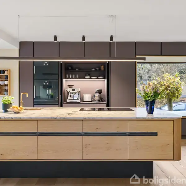 Et moderne køkken med en central køkkenø i træ udstyret med en stilfuld vandhane og friske blomster. I baggrunden ses indbygget kaffemaskine og hylder. Store vinduer giver lys.