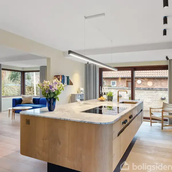 Køkken med træ-ø og sort kogeplade, blomsterdekor på bordet, rummeligt opholdsrum med blå sofa i baggrunden, store vinduer og udsigt til murstensbygning og have.