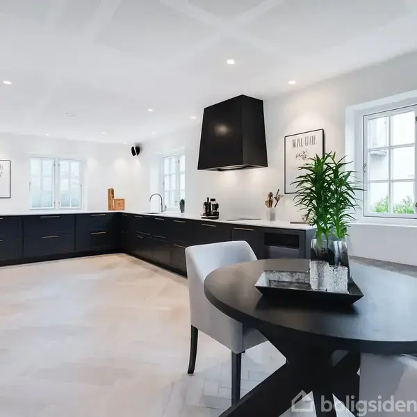 Et moderne køkken har sorte kabinetter langs væggene. En rund sort spisebord står ved et vindue, med en plante og bakke ovenpå. Rummet er lyst og rummeligt.